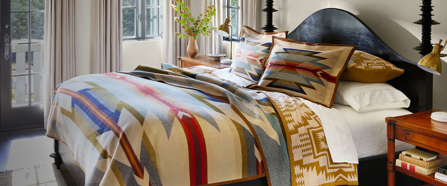 Bed with blankets quilts and pillows stacked in a comfortable sunlit room.